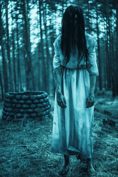 Dziewczyna z długimi czarnymi włosami w obrazie strasznego ducha zombie stoi wśród ciemnego lasu na tle kamienia dobrze. - Zdjęcie, obraz