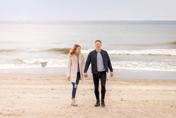 Attractive young couple walking along the shore of a sandy beach, on a spring romantic holiday, outdoors. Travel tourists lifestyle. Couple enjoying loving time together. - Photo, image