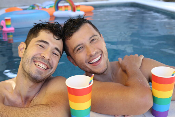 ευτυχισμένο ζευγάρι ομοφυλόφιλων που απολαμβάνουν το χρόνο τους μαζί στην πισίνα με χάρτινα φλιτζάνια σε χρώματα της σημαίας Igbtq - Φωτογραφία, εικόνα