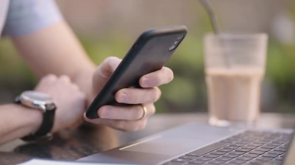 Primer plano un hombre con un teléfono móvil se dedica a la banca en línea, compra en Internet y escribe mensajes en las redes sociales mientras está sentado con un ordenador portátil - Imágenes, Vídeo