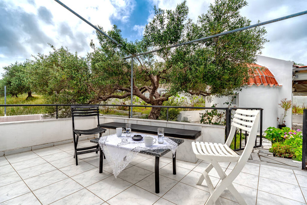 Backyard βεράντα ενός εξοχικού μεσογειακού σπιτιού με τραπέζι και δύο καρέκλες μαύρο και άσπρο. Μικρό τραπέζι σερβιρισμένο με cun καφέ και ποτήρια νερού, στον κήπο με τις ελιές - Φωτογραφία, εικόνα
