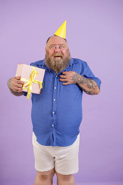 Překvapený muž s nadváhou v těsné modré košili drží dárkové krabice pózovat na fialovém pozadí - Fotografie, Obrázek