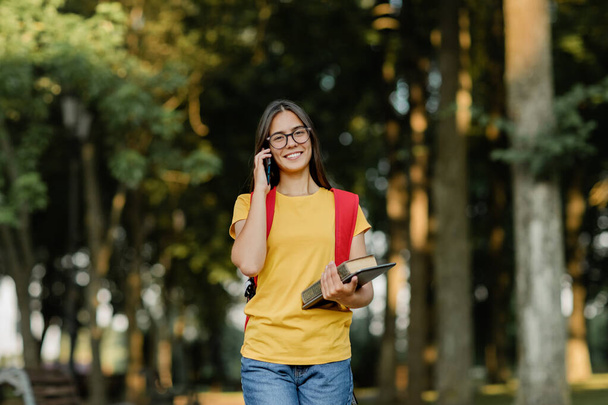 Portrait of a beautiful brunette with glasses and a backpack talking on the phone in the park - Photo, Image