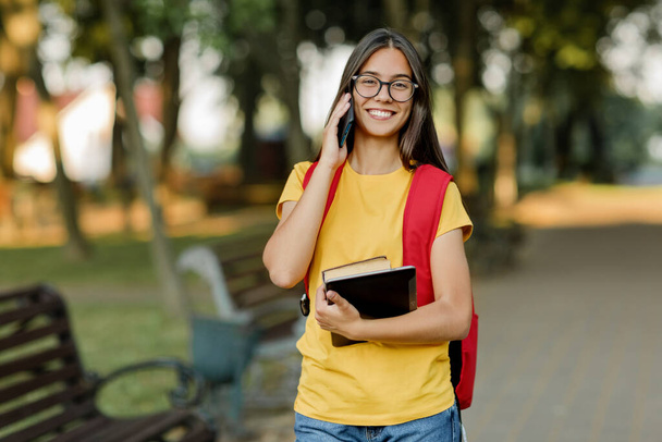Portrét krásné brunetky s brýlemi a batohem mluvící po telefonu v parku - Fotografie, Obrázek