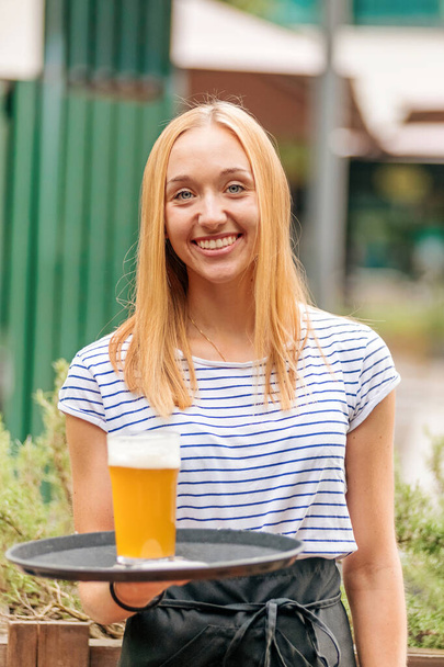 Ευτυχισμένη φιλική νεαρή σερβιτόρα παραδίδει ένα ποτήρι κρύα μπύρα σε ένα δίσκο με ένα ακτινοβόλο χαμόγελο σε ένα υπαίθριο εστιατόριο ή παμπ - Φωτογραφία, εικόνα
