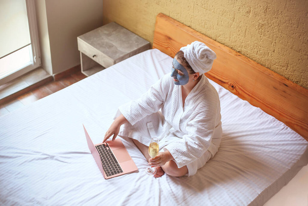 Mavi kozmetik maskeli genç hippi kadın evde yatağında oturmuş bilgisayarla şampanya içerken şampanya içerken gülümsüyor. Üst görünüm - Fotoğraf, Görsel