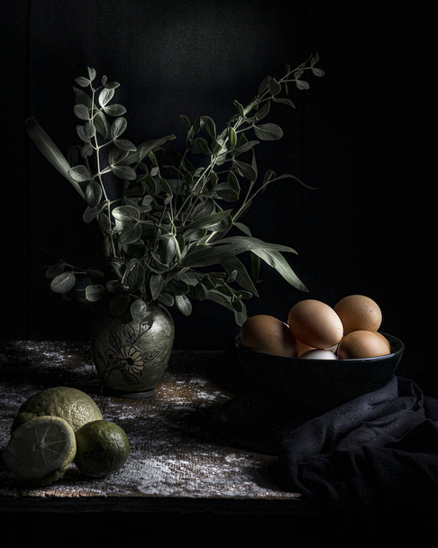 A still life of eggs in a bowl, lemons, and a plant in a vase on a wooden table - Фото, изображение