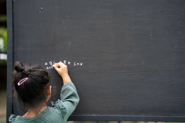 Вид сзади на милую маленькую тайскую девочку, пишущую алфавит на доске - Фото, изображение