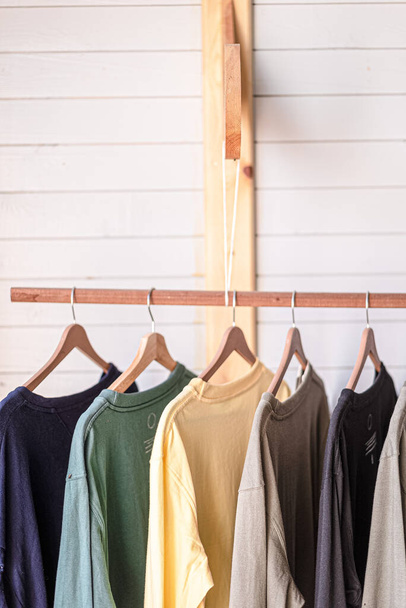 A view of a couple of colorful t-shirts with hangers hanging on the wooden pole in the room - Фото, зображення