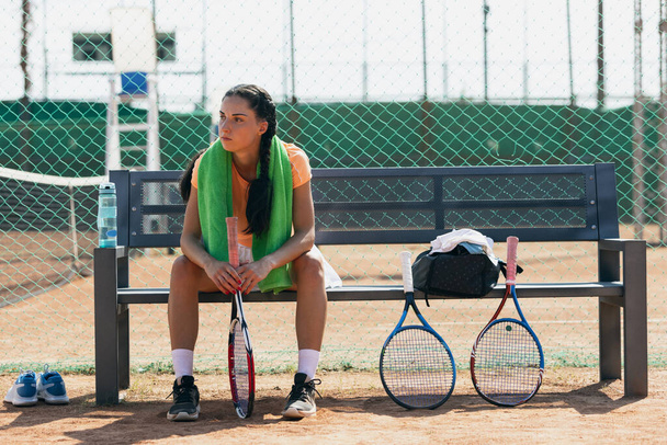 Front image of young sportswoman sitting and resting on bench on tennis court - Photo, Image