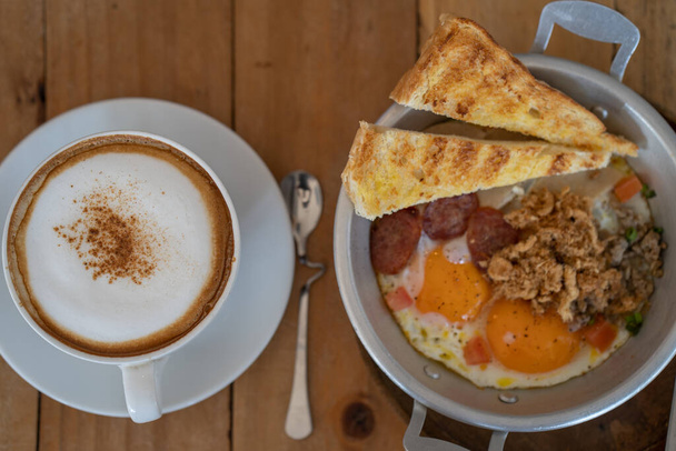 A top view of breakfast with fried eggs and toasts and a cup of cappuccino on a wooden table - Foto, afbeelding