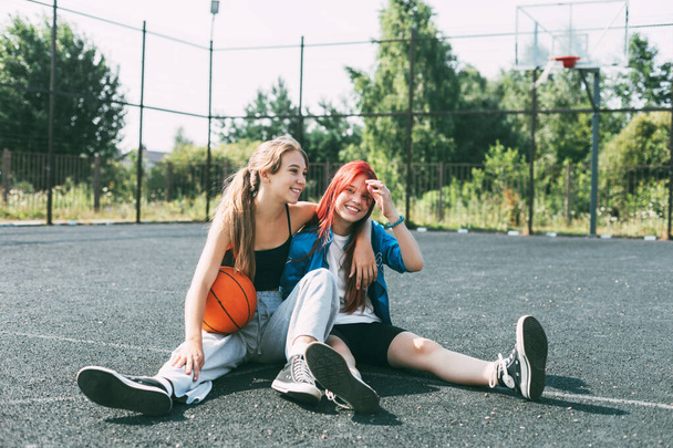 Two girls in sports clothes and with a basketball are chatting, sitting on the playground. Sports, competition, friendship - Photo, Image