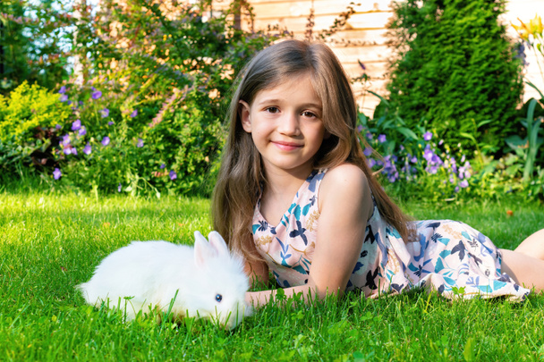 Tyttö leikkii kotimaisen valkoisen kanin kanssa puutarhan nurmikolla kesällä. Onnellinen lapsuus käsite. Lemmikkieläinten ja lasten ystävyyden käsite. Kani syö ruohoa luonnossa kesäpäivänä.. - Valokuva, kuva