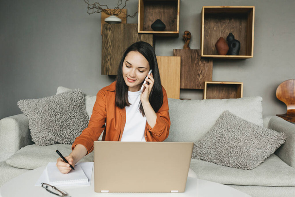 Eine schöne junge Frau in lässiger Kleidung benutzt Laptop und Handy, während sie zu Hause arbeitet. Eine Frau schreibt mit einem Stift auf Papier. - Foto, Bild