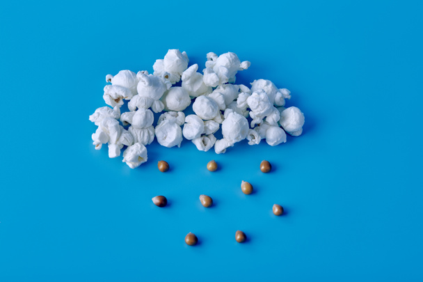Palomitas de maíz de aire blanco en forma de nube de la que gotea la lluvia, gotas de semillas de maíz sobre un fondo azul - Foto, Imagen