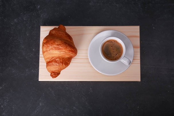 Fresh baked croissant on chopping board, wooden pad, white mug of expresso on dark concrete table. Cup of hot coffee, buns, rolls close up. Food, French breakfast, morning menu, cafe concept. Top view - Фото, изображение