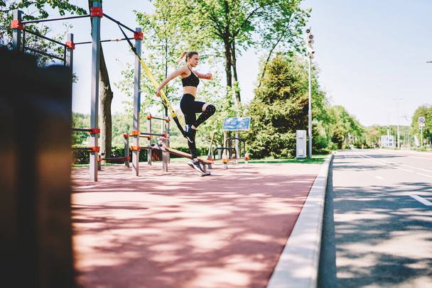 Slim young athletic female with blonde hair in black sportswear training with yellow elastic band in sports ground with fitness equipment in street in summer - Photo, image