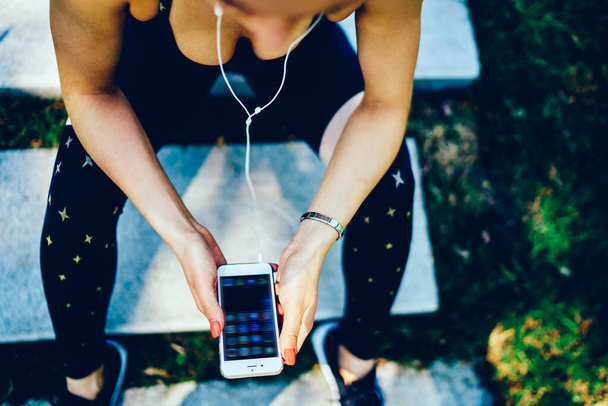 From above of crop active lady in sportswear with earphones browsing phone checking social media relaxing from physical exercise sitting on stairs in park - Foto, Imagem