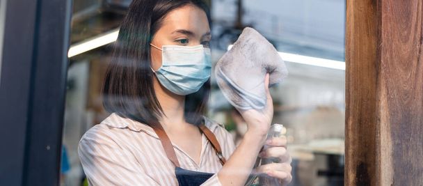 Caucasian attractive waitress wear protective mask to prevent from Covid-19 pandemic at cafe. Young business owner girl barista in apron clean mirror with disinfectant and wipe before welcome customer - Zdjęcie, obraz