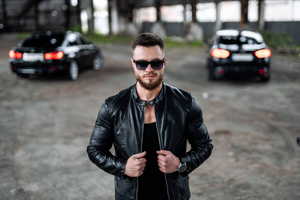 Stylish handsome man with sunglasses in fashionable clothes standing with the luxury black cars on the background. Closeup - 写真・画像