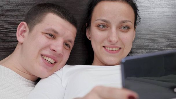 Молода пара, що тримає смартфон в руці, фотографується разом, знімає відеоблог на ліжку. Щаслива сім'я чоловік і дружина фотографують онлайн, використовуючи сучасний мобільний телефон вдома на дивані в спальні
. - Фото, зображення