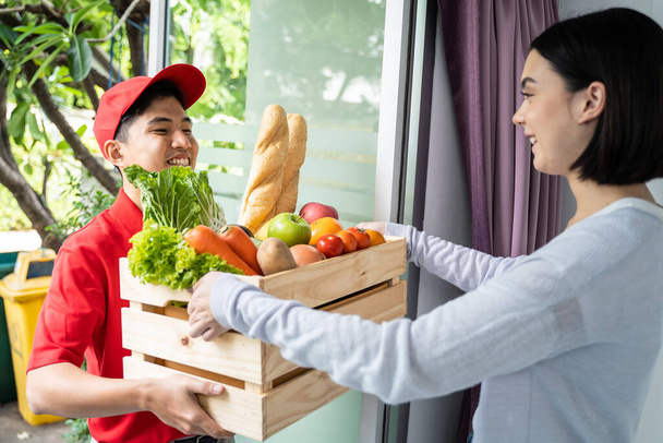 Ενεργός άνθρωπος παράδοση τροφίμων που εργάζονται με κόκκινη στολή παραδώσει κουτί φρούτων και ψωμιού σε νεαρή κοπέλα πελάτη. Όμορφη κυρία με χαμόγελο πρόσωπο λήψη κουτί τροφίμων από αγγελιοφόρος μπροστά από την πόρτα στο σπίτι - Φωτογραφία, εικόνα