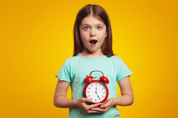 Shocked girl in blue t shirt holding alarm clock isolated over yellow wall - Φωτογραφία, εικόνα