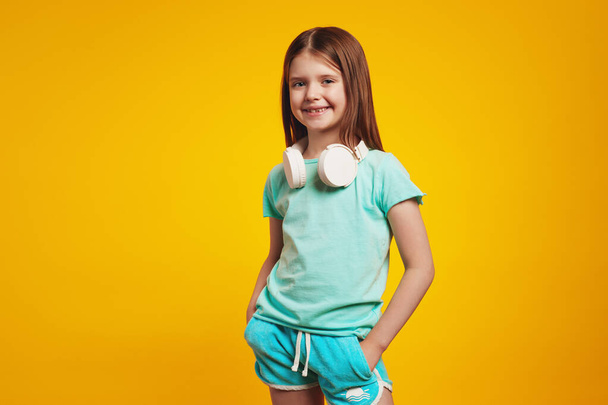 Little cute girl wearing white headphones on neck, keeping hands in pockets - Fotografie, Obrázek