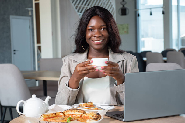 Αφροαμερικανή γυναίκα με κοστούμι επιχειρήσεων με φορητό υπολογιστή και πίτσα πίνοντας τσάι στο καφέ - Φωτογραφία, εικόνα