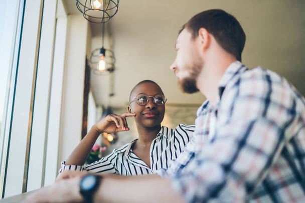 Concentrated African American woman in glasses attentively listening to relaxed man sitting at table before window in light designed cafe - Foto, afbeelding