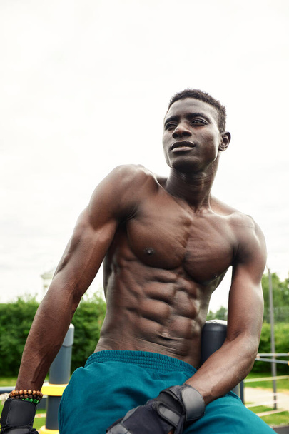 Muscular young man exercising at the sports field. African man looking to the side while doing horizontal bar exercises. Shirtless male model exercising outdoors. - Фото, зображення