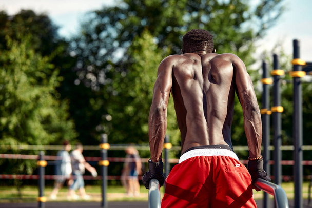 Svalnatý africký americký atlet s nahým vymodelovaným trupem dělá kliky na nerovných tyčích v městské krajině, Záběr zezadu, kopírovací prostor - Fotografie, Obrázek