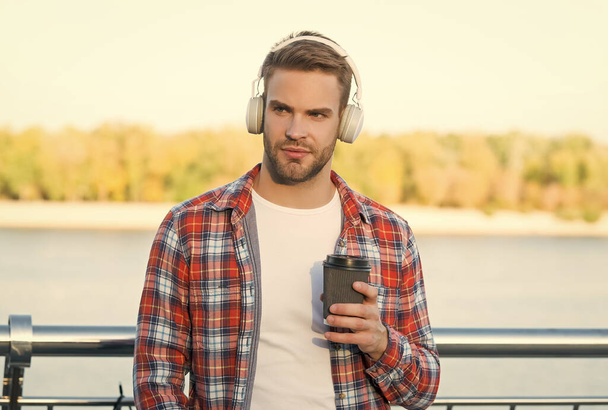 serious bearded man listen music in digital earphones and drinking coffee to go from paper cup wearing checkered shirt outdoor, good morning - Photo, image