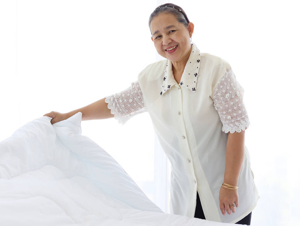 Happy souriant asiatique femme âgée femme au foyer nettoyage et faire le lit dans la chambre, grand-mère faire le ménage et le nettoyage, vieille femme de chambre changeant la couverture blanche et drap. - Photo, image