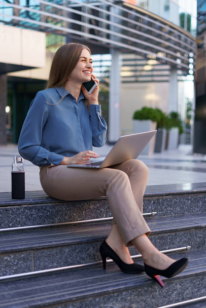 Kobieta rozmawiająca przez telefon komórkowy i pracująca z laptopem siedząc na schodach - Zdjęcie, obraz