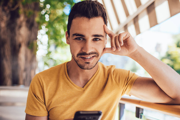 Young content male hipster in casual yellow t shirt surfing on mobile phone while smiling and looking at camera at outdoors terrace of cafe  - Zdjęcie, obraz