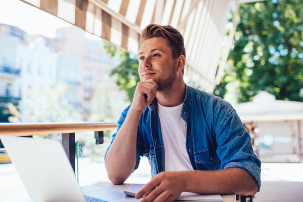 Young prosperous male freelancer sitting on cafe terrace with laptop computer thinking about future vacation, creative man blogger concentrated on article publication looking away during distance wor - Φωτογραφία, εικόνα