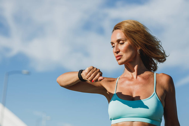 Portrait of a middle age fit woman looking on smartwatch and resting after hard training outdoor. - Fotoğraf, Görsel