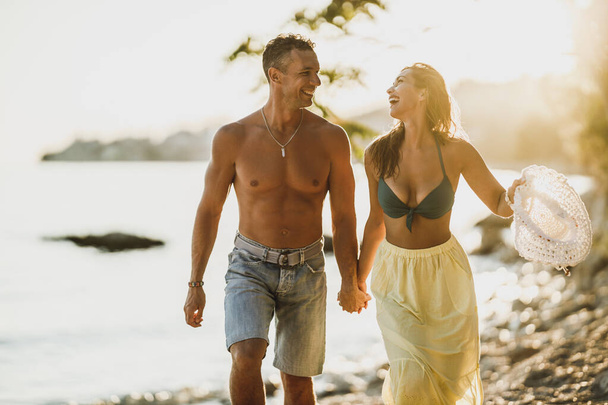 Glückliches Paar hält Händchen, geht spazieren und fühlt sich verspielt, während es den Sommertag am Ufer verbringt. - Foto, Bild