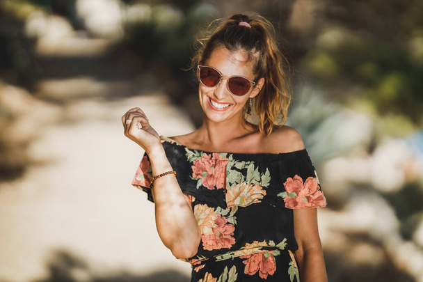 Portrait d'une jeune femme souriante avec des lunettes de soleil tout en marchant dans une pinède près de la mer. - Photo, image