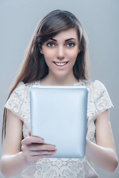 Una giovane bella donna sorridente con i capelli lunghi scuri sta tenendo una tavoletta d'argento. Su uno sfondo grigio. - Foto, immagini