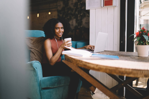 Curly haired cheerful ethnic woman in casual clothes drinking coffee surfing on laptop sitting in blue sofa in outdoor terrace  cafe in summe - 写真・画像