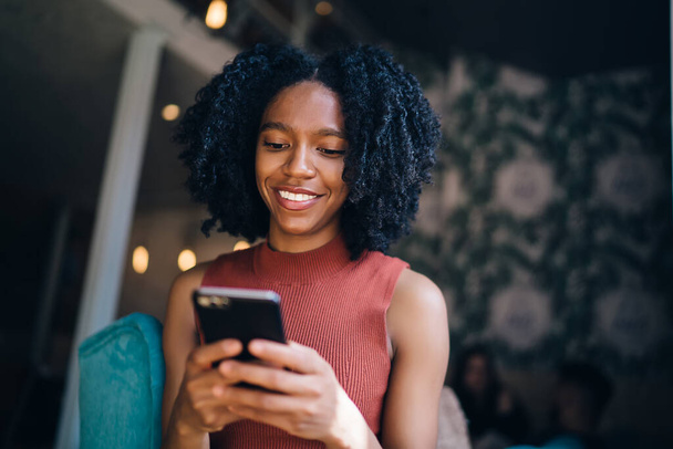 African American cheerful young woman with Afro hairstyle in casual clothes using phone surfing social media relaxing in modern cafe - Fotoğraf, Görsel