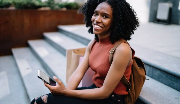 Excited black curly haired young woman in casual wear using smartphone resting sitting on stairs in street looking at camera - Foto, afbeelding