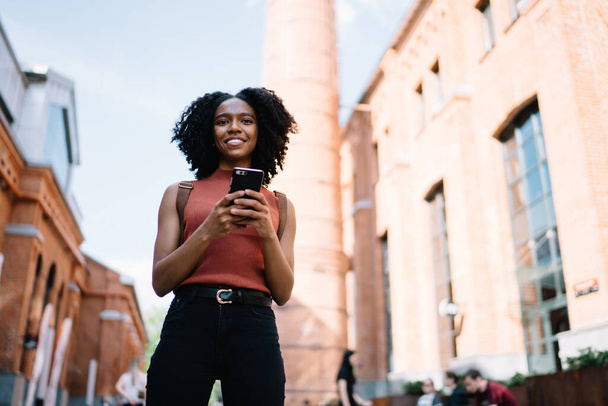 View from below of positive smiling black curly haired woman in casual wear using phone chatting with friend strolling in street looking at camera - 写真・画像