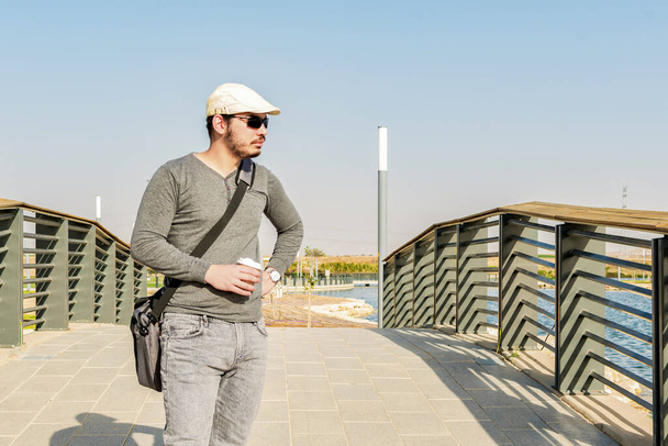 A young Israeli man with a cap and sunglasses taking a walk on a bridge at a sunny park outside - 写真・画像