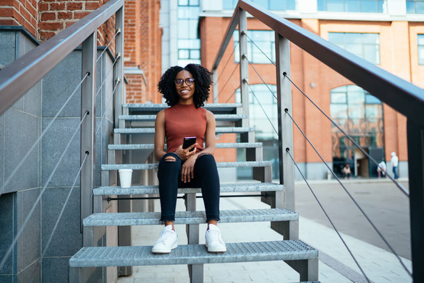 Young content African American spectacled female dressed in casual clothes sitting on stair in street with mobile phone at daytime and looking at camera with smile - Zdjęcie, obraz