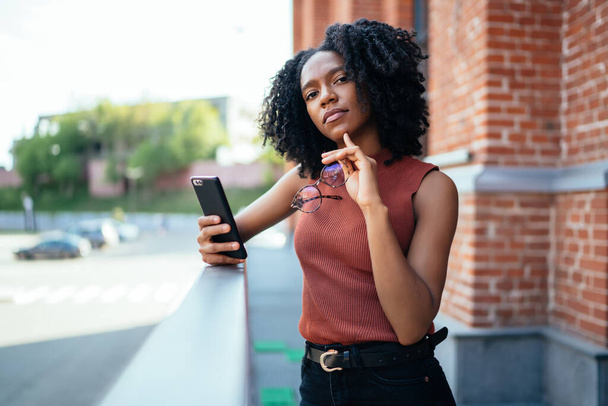 Young proud black woman holding cellphone and glasses while standing over fence next to brick building and looking at camera - Photo, Image