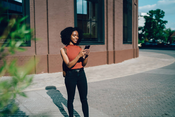 Confident pleased African American female student standing and chatting in social media on smartphone on background with modern university campus - Foto, Imagem