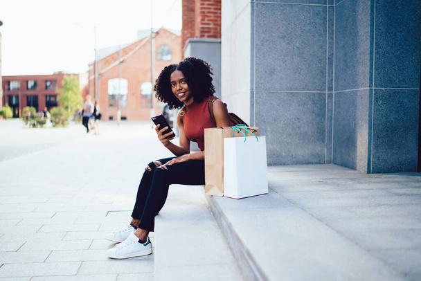 Cheerful content African American curly haired woman sitting on stairs with paper bags in sleeveless casual shirt texting on mobile phone in street in summer - Foto, immagini